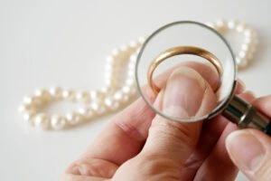 A hand holds gold wedding band , shown through a magnifying glass, with a pearl necklace in the background